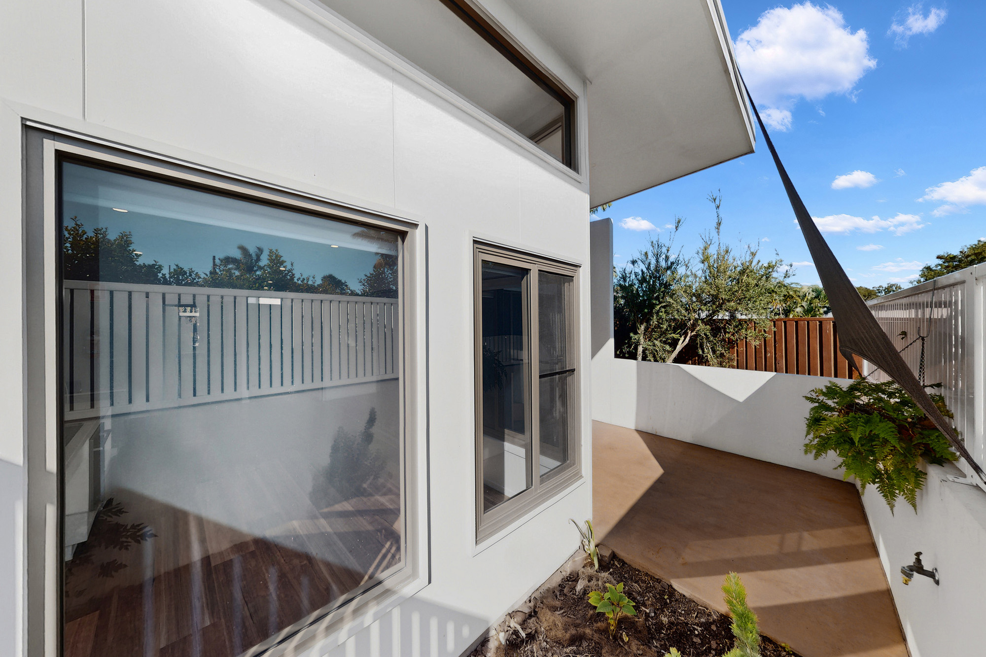 Modern white exterior of a building with large windows, a small patio area, plants, and a shade sail. The sky is blue with a few clouds and trees are visible beyond the fence.