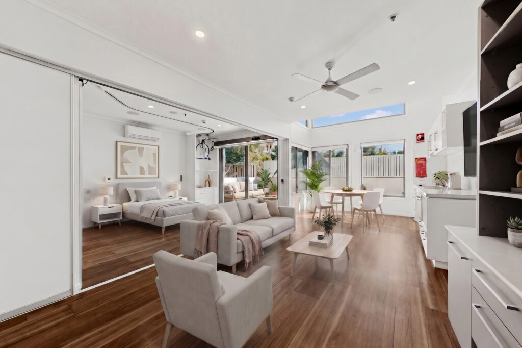 A modern, open-plan studio apartment with a living area, bed, dining table, and kitchen. Large windows let in natural light, and the room features wood floors, white walls, and minimalist decor.