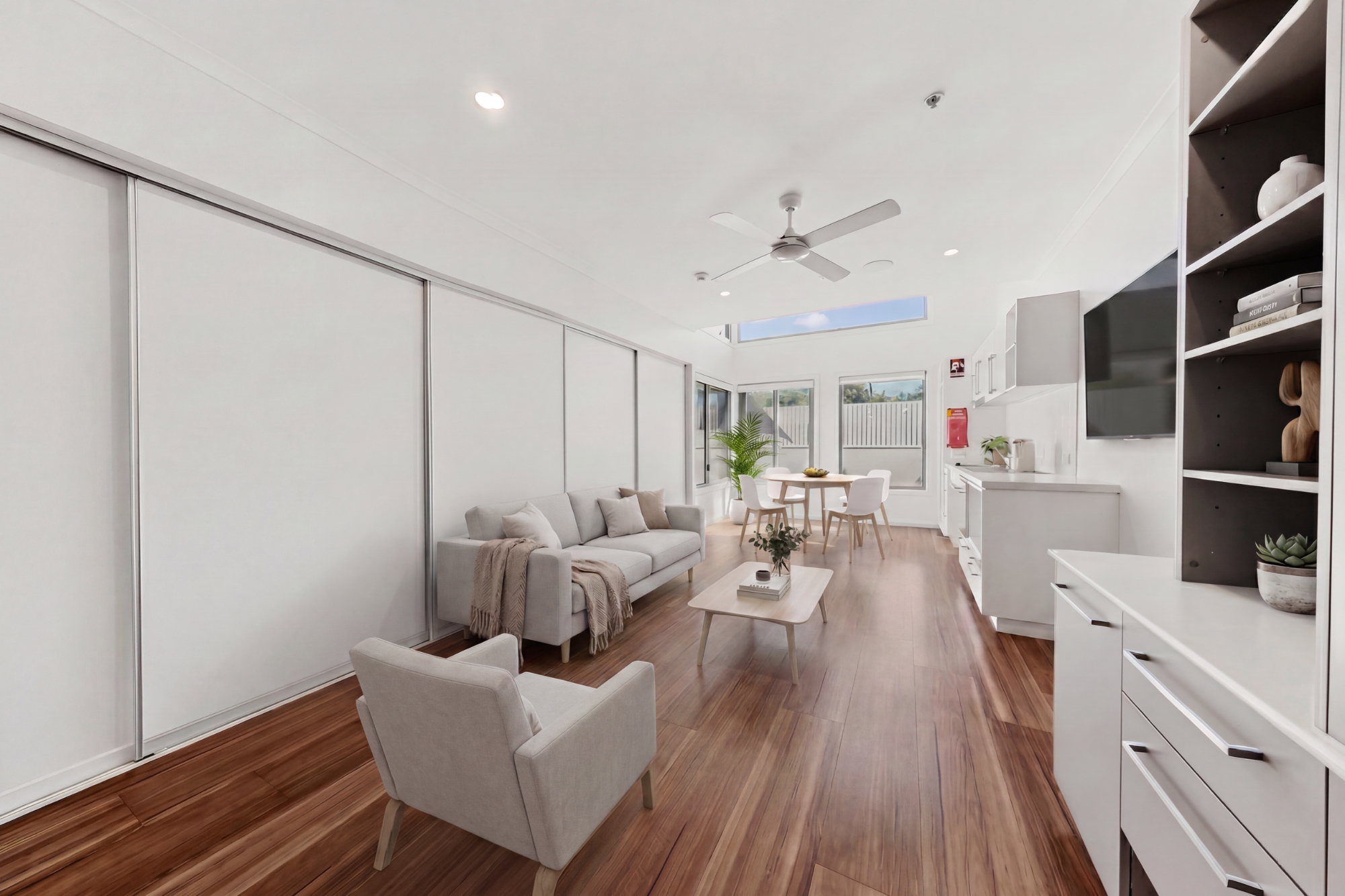 A modern, bright living room with wood flooring, white walls, a gray sofa, armchair, coffee table, TV, built-in shelves, kitchen area, and large sliding glass doors letting in natural light.