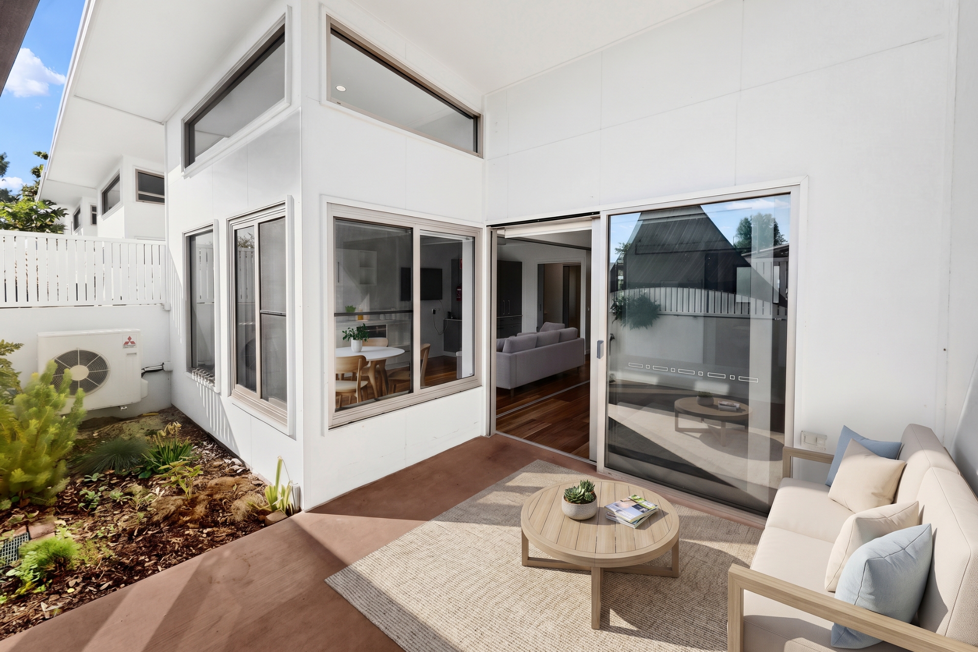 A modern patio with a beige outdoor sofa and round coffee table on a rug, next to sliding glass doors leading to a bright living and dining area. Large windows overlook a small garden with plants.