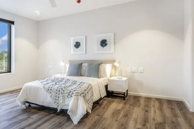 Modern bedroom with wooden floors, a double bed with white and gray bedding, two gray pillows, two bedside tables with lamps, and two framed abstract artworks on the wall above the bed.