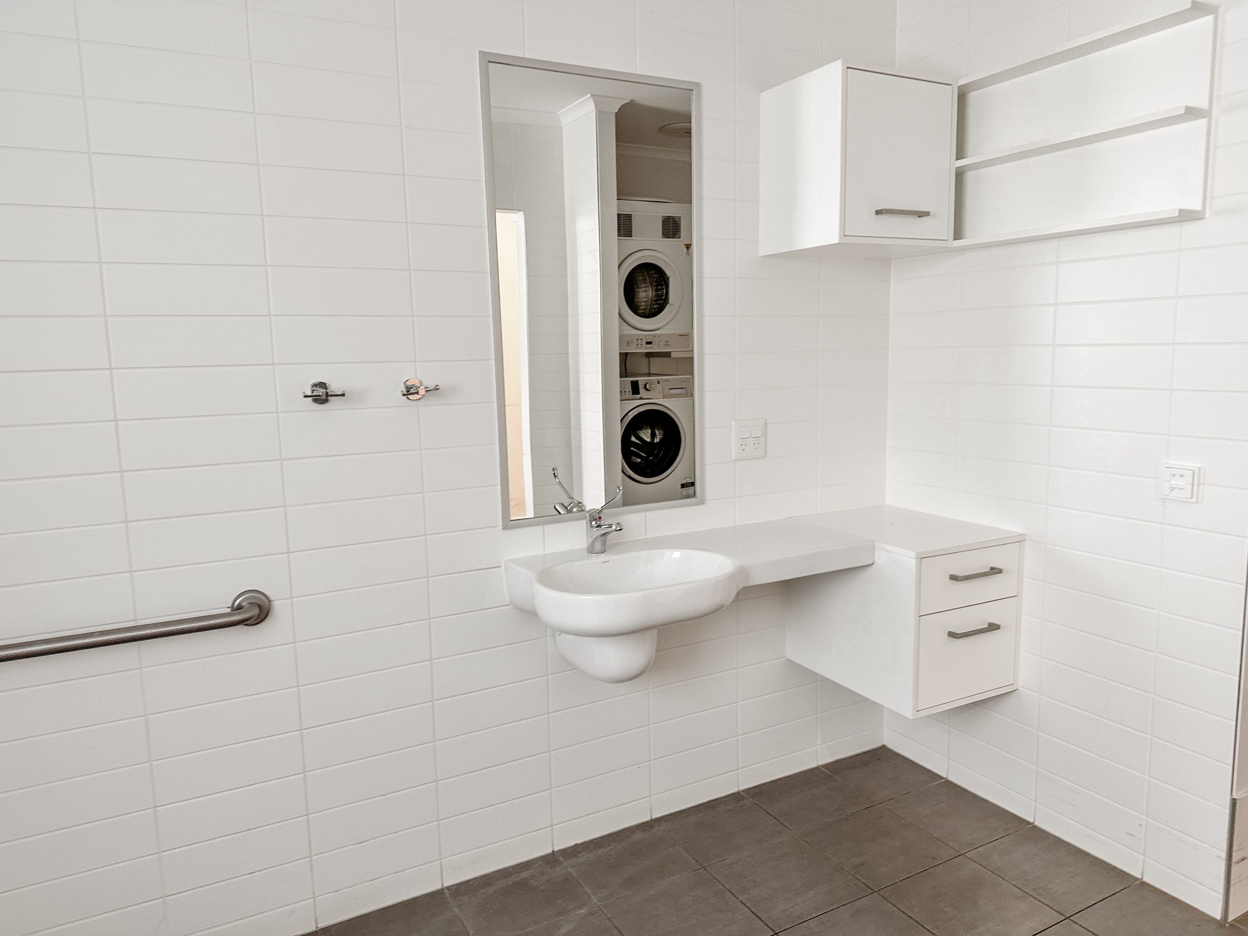 A modern white bathroom with a wall-mounted sink, cabinets, towel rack, and a mirror reflecting a stacked washer and dryer. The walls are tiled and the floor has large gray tiles.