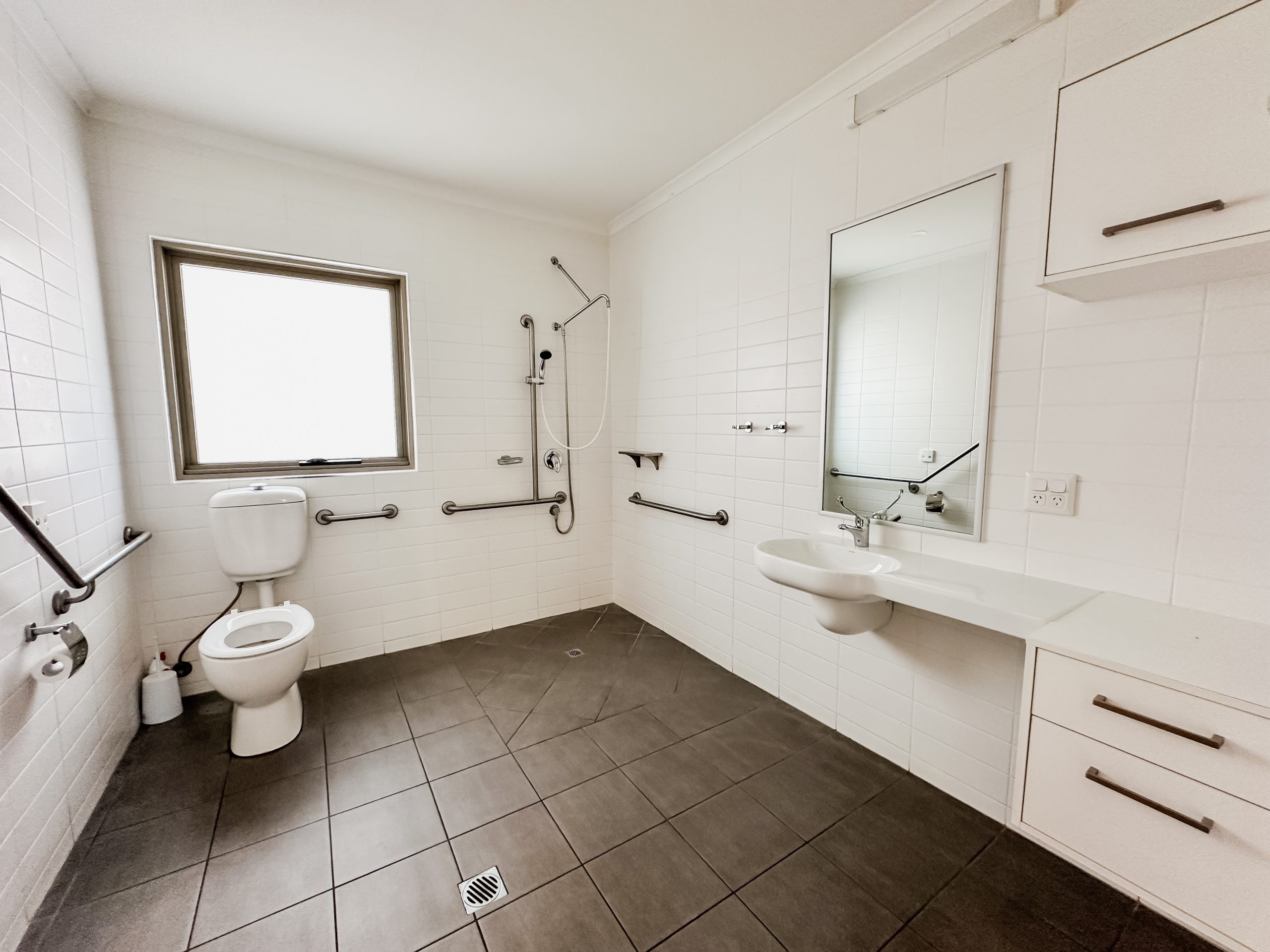 Accessible bathroom with grab bars, a toilet, a roll-in shower with handheld showerhead, a wall-mounted sink, a mirror, tiled floors, white walls, and a frosted window letting in natural light.