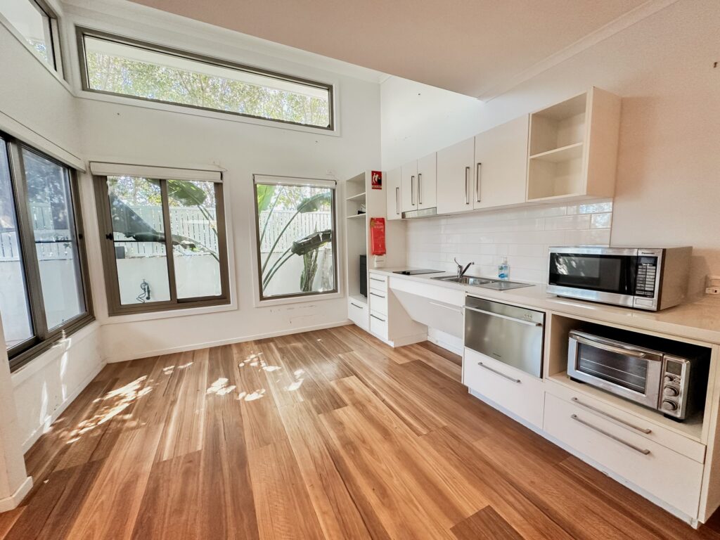 Modern kitchen with wooden floors, white cabinets, large windows letting in natural light, a stainless steel sink, microwave, and drawers. Greenery and a fence are visible outside through the windows.