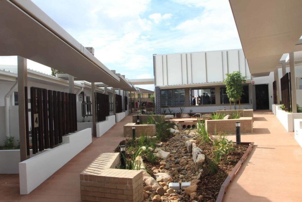 A modern outdoor courtyard with landscaped garden beds, a rocky dry creek, trees, and two single-story buildings featuring large windows and shaded walkways. The sky is partly cloudy.