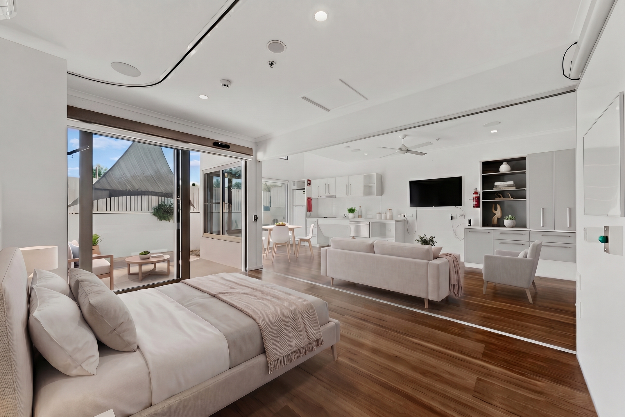 Modern open-plan studio apartment with a bed, living area, and kitchen. Large glass doors lead to a patio with outdoor seating. Space is bright with white walls, wood flooring, and neutral-toned furniture.
