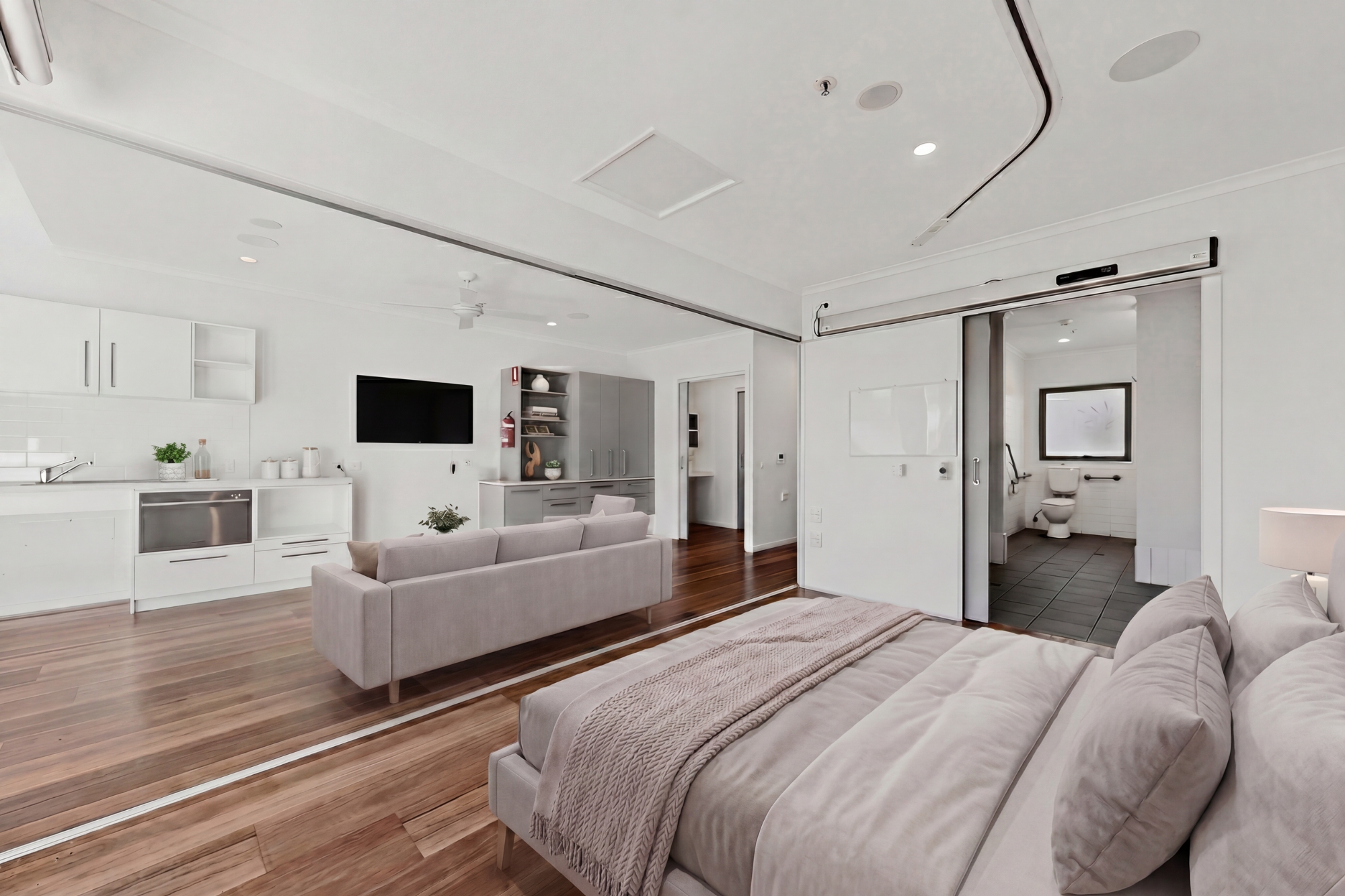 Modern open-plan studio apartment with a bed, sofa, wall-mounted TV, kitchenette, and a sliding door leading to a bathroom. The room features white walls, wooden floors, and contemporary furnishings.
