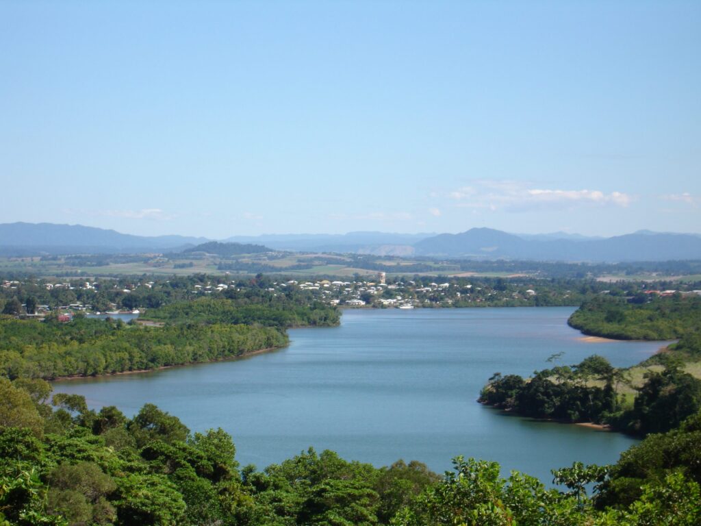 A wide river curves through lush green vegetation with a small town on its banks, set against distant hills and a clear blue sky on a sunny day.