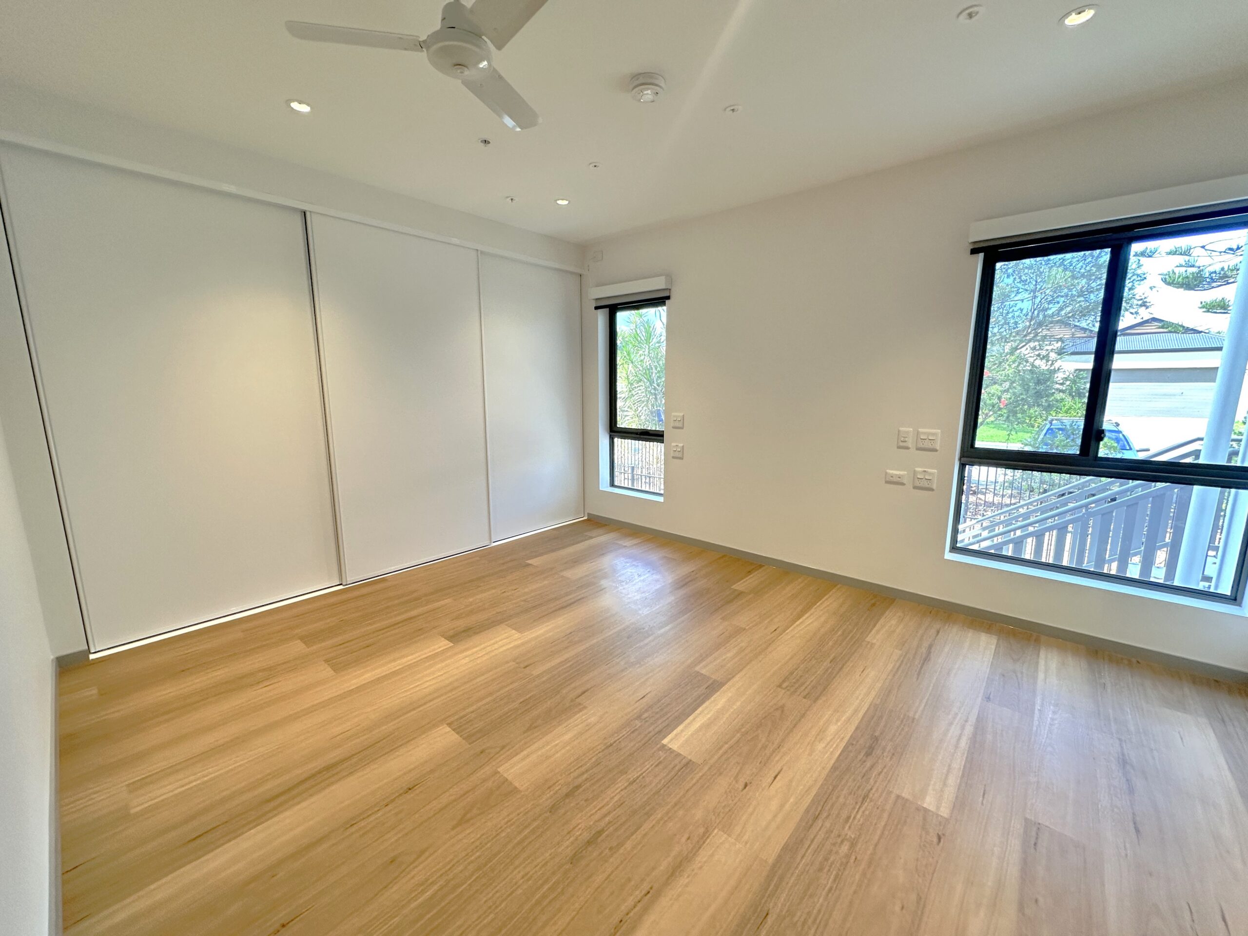 Bright, empty room with light wooden floors, white walls, a ceiling fan, built-in white closets, and two large windows letting in natural light, showing trees and a deck outside.