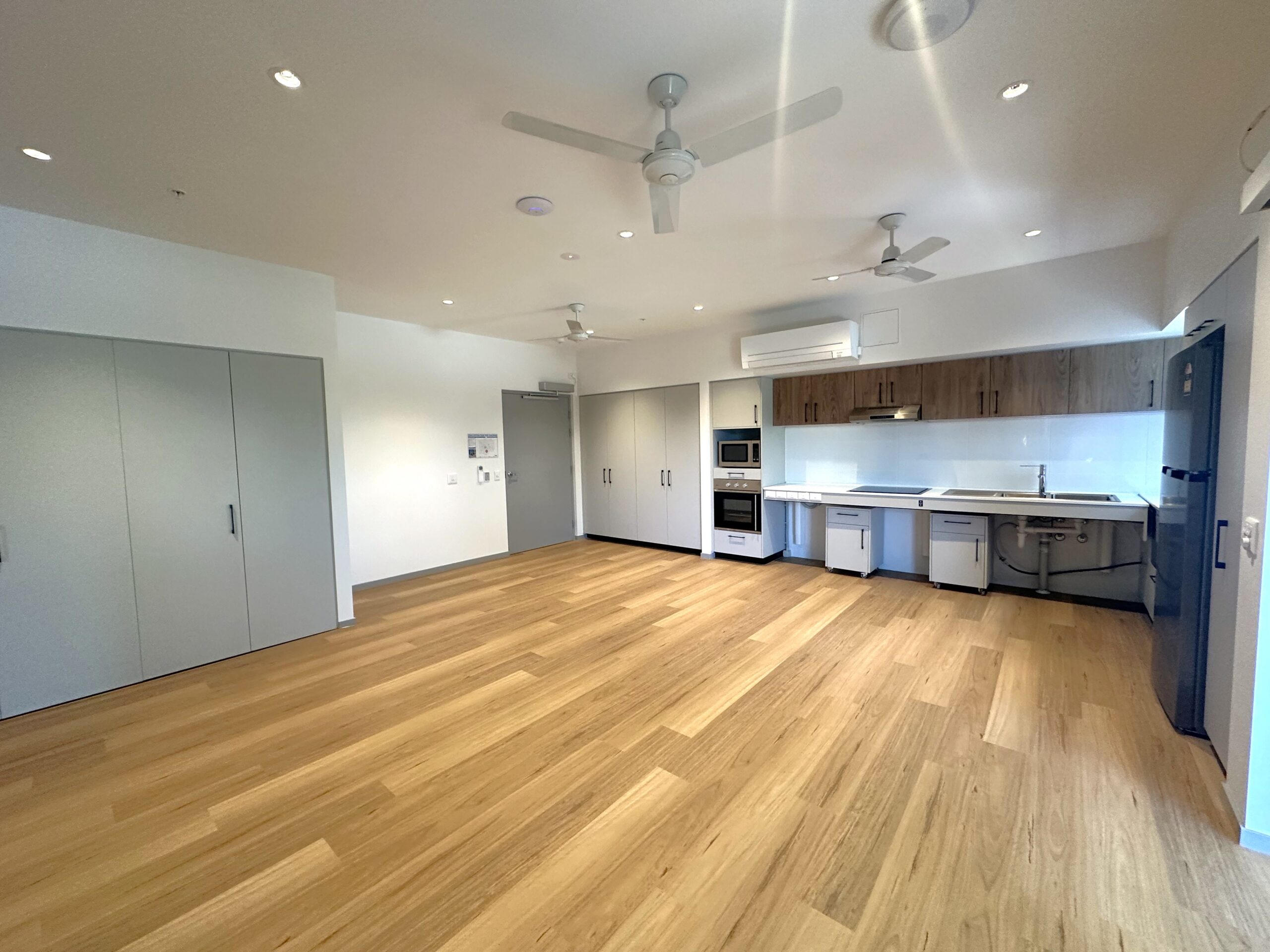 Modern, empty apartment with light wood floors, white walls, a built-in kitchen featuring microwave, oven, sink, and wooden cabinets. There are ceiling fans, cupboards, and lots of natural light.
