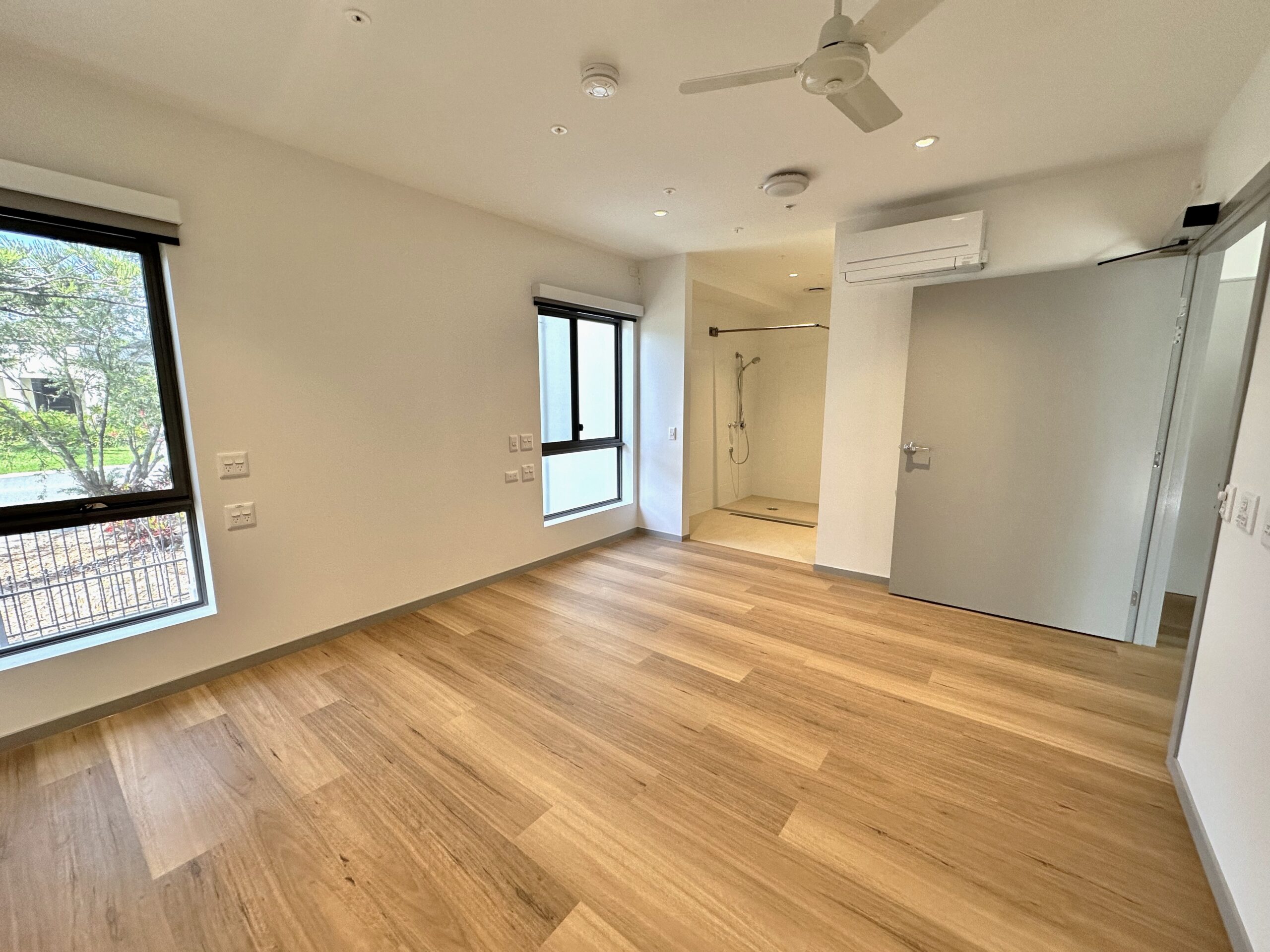 A bright, unfurnished room with light wood floors, two windows, a ceiling fan, an open door, and an accessible en suite bathroom with a walk-in shower. White walls and modern fixtures are visible.