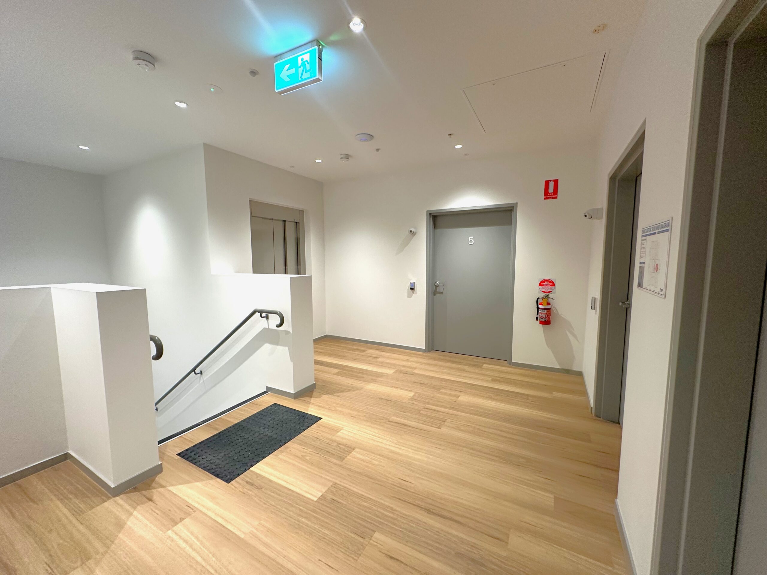 A brightly lit hallway with light wood flooring, white walls, several doors, a fire extinguisher, an emergency exit sign, a stairway with a railing, and a black floor mat near the steps.