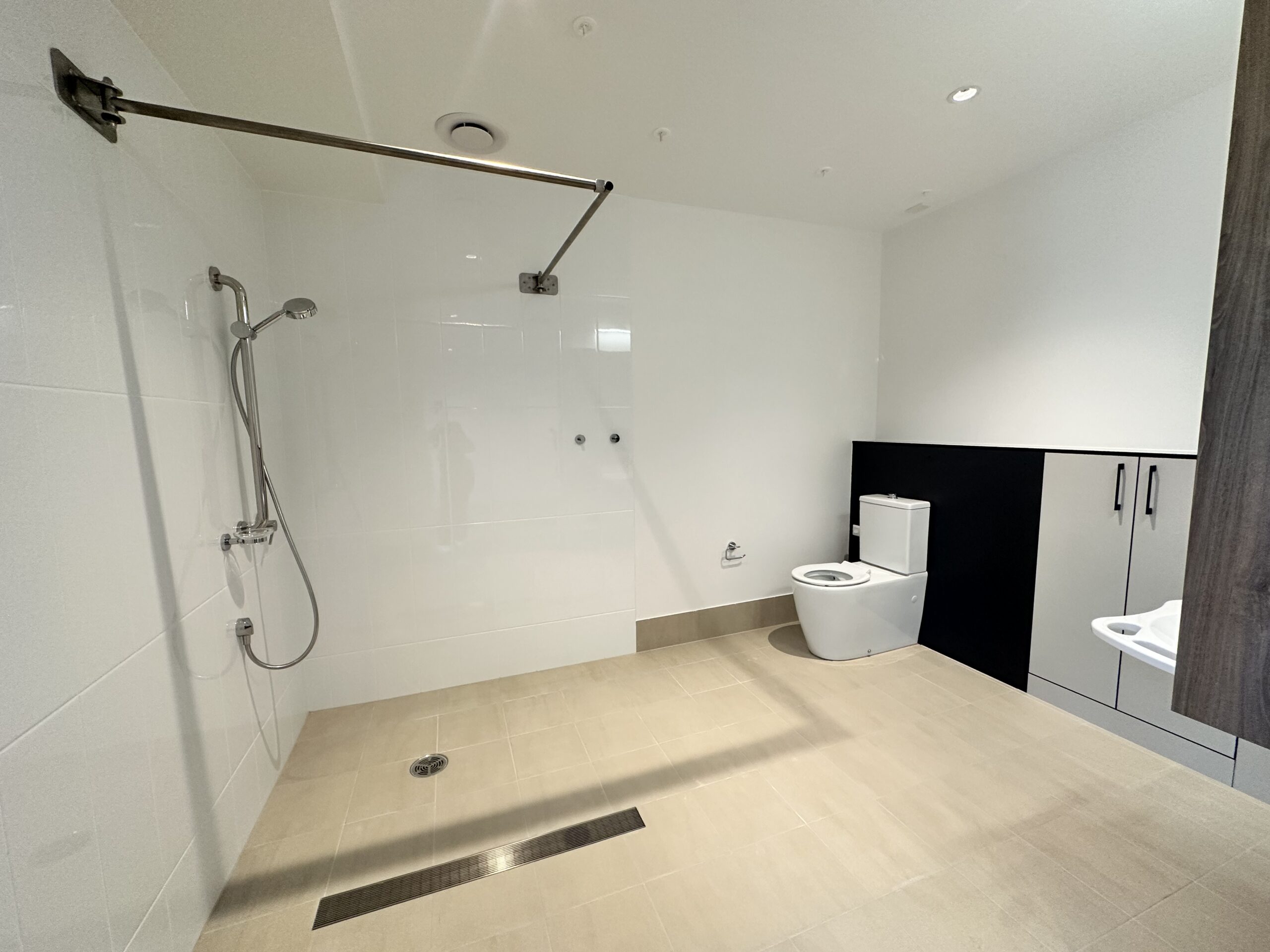 A spacious accessible bathroom with a walk-in shower, wall-mounted showerhead, floor drain, toilet, and built-in cabinets. The room has white walls, beige tiles, and bright lighting.
