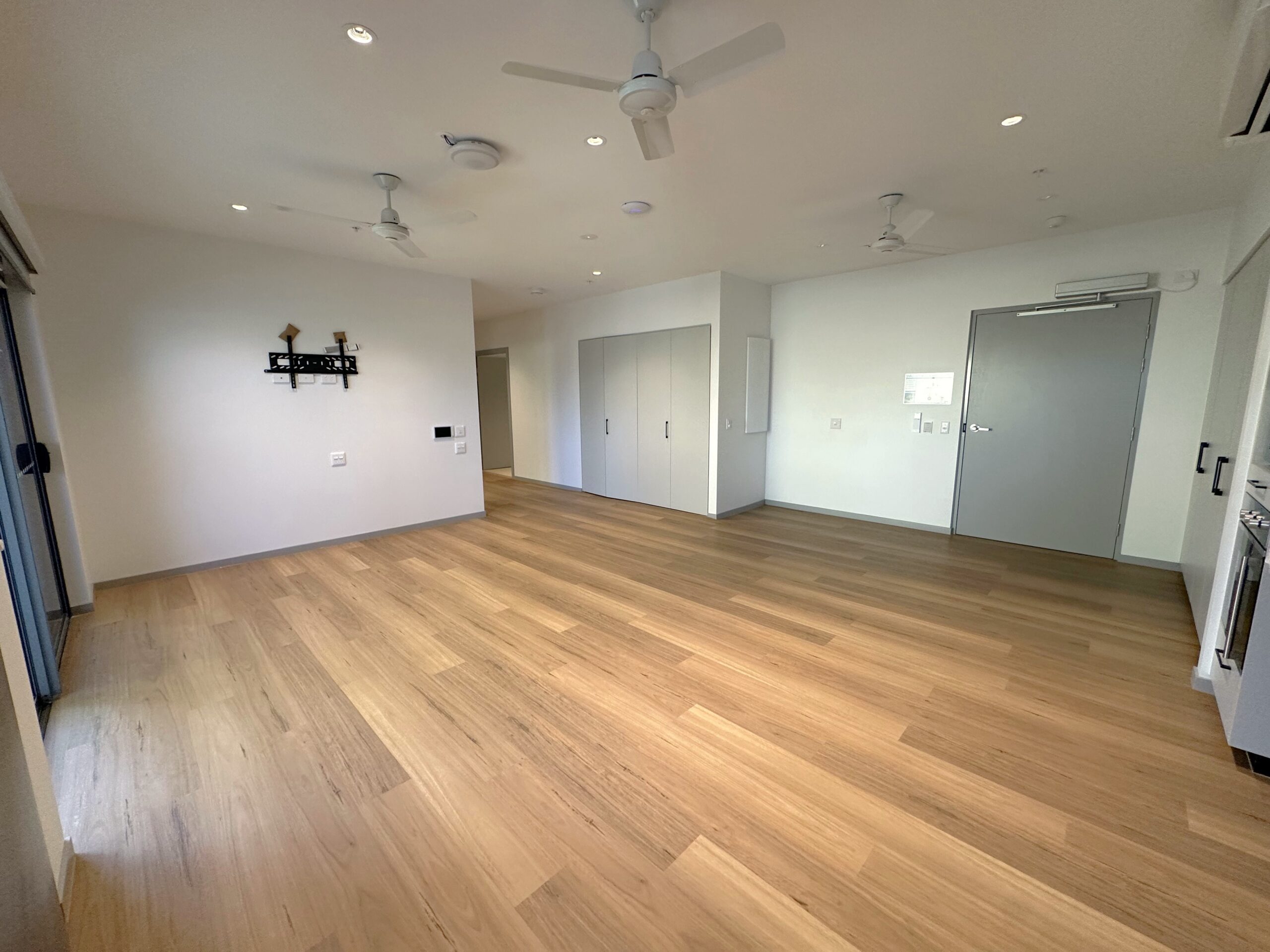 A spacious, empty room with light wood flooring, white walls, multiple ceiling fans, and built-in closets. There are large windows on one side, a grey door, and a black wall-mounted shelf.