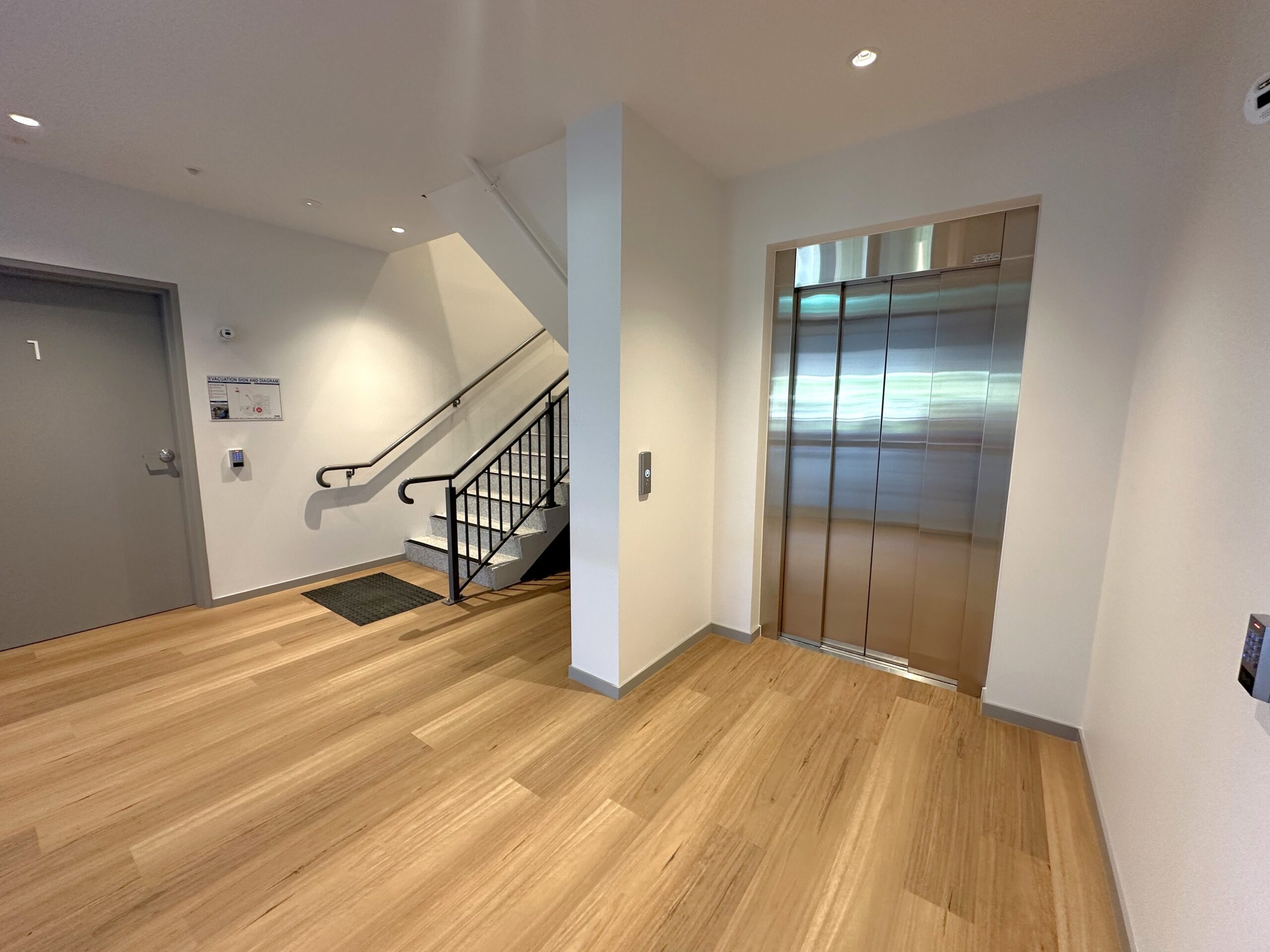 A modern building hallway with light wood floors, a closed elevator with stainless steel doors on the right, a door labeled 