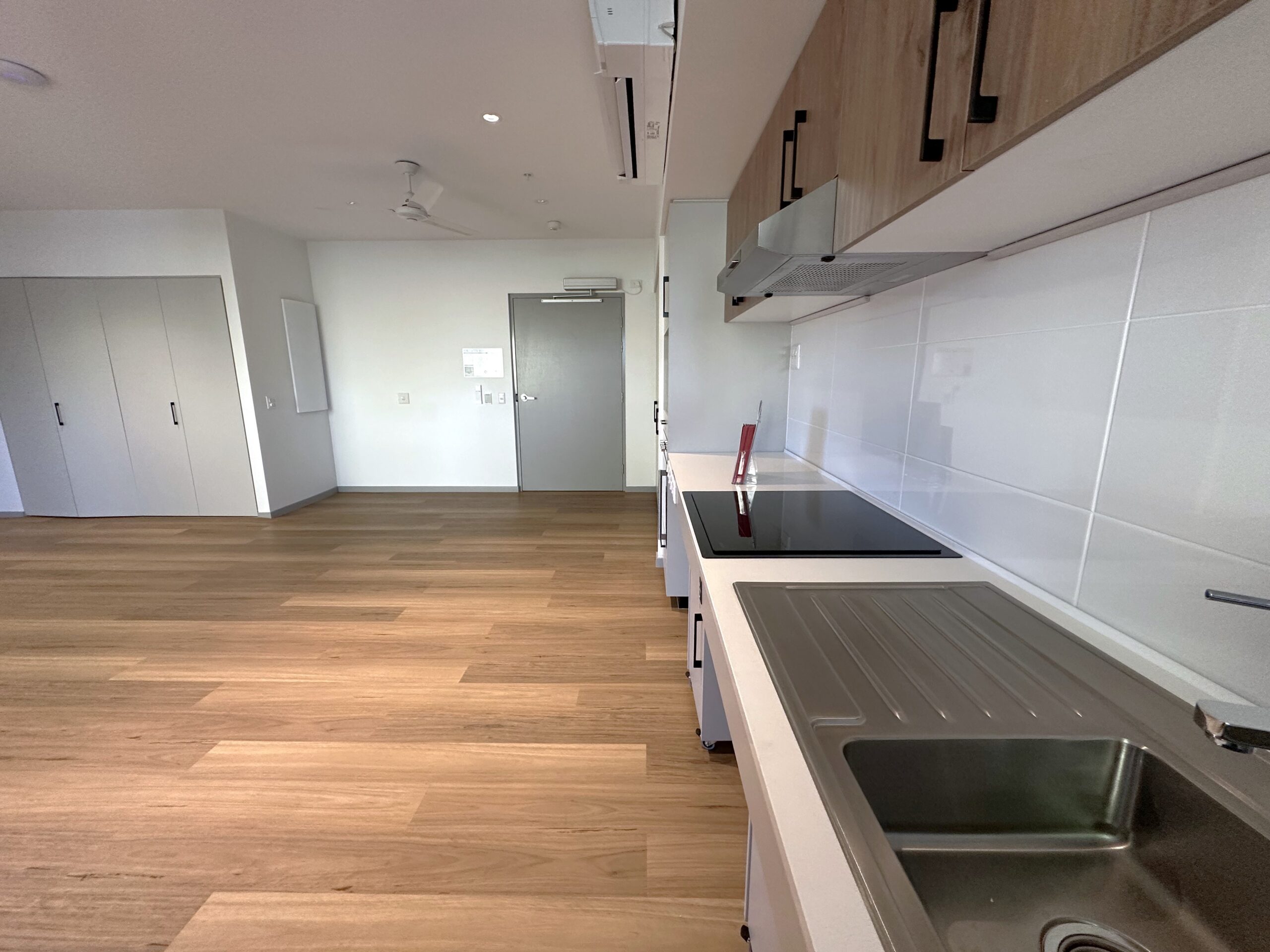 Modern apartment kitchen with wood flooring, white cabinets, a stainless steel sink, electric stovetop, and white tile backsplash. An open living space and built-in closets are visible in the background.