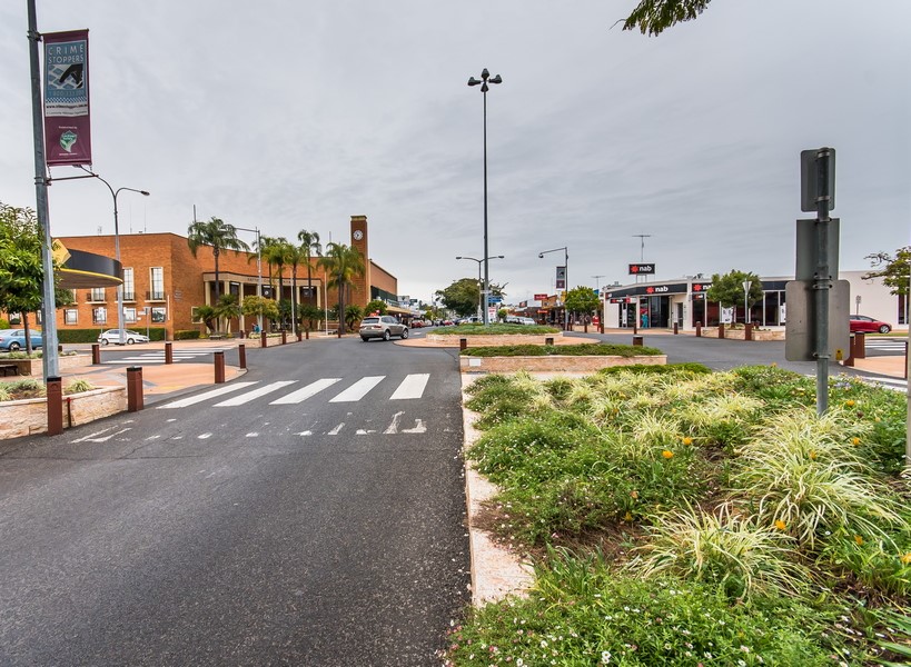 A crosswalk and road run through a small town center with shops, greenery, streetlights, and a clock tower under a cloudy sky. Cars are parked along the sides, and banners hang from lamp posts.