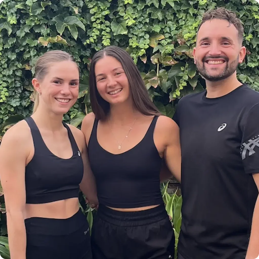 Three people smile and pose together outdoors in front of leafy green foliage. They are wearing black athletic wear and appear happy and relaxed.