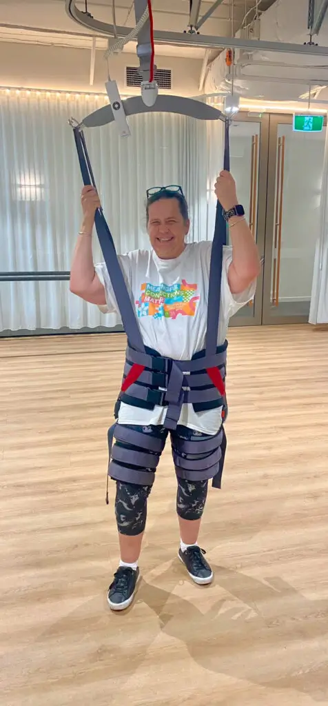 A person standing indoors, smiling while wearing a harness lift system. They are dressed in a colorful t-shirt, black patterned leggings, and sneakers, and holding onto the support straps with both hands.