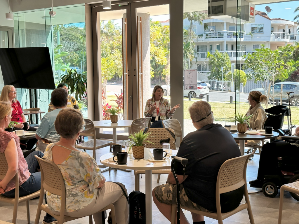 A group of people sit around tables in a bright, modern room with large windows, attentively listening to a woman speaking at the front. There are plants on the tables and outdoor greenery is visible through the windows.
