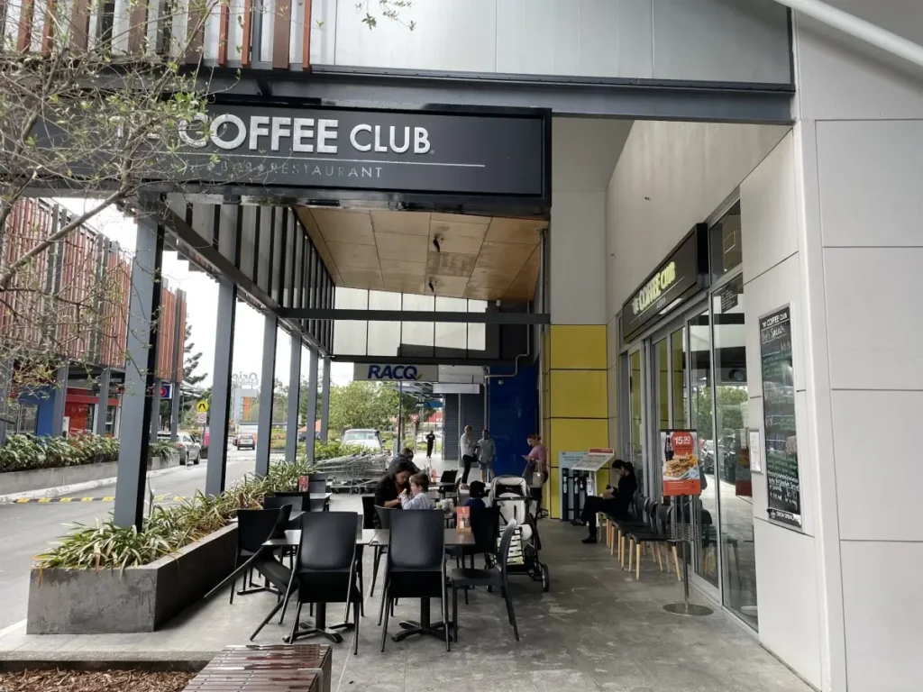 Outdoor seating area of The Coffee Club café with several people sitting at tables. The café is under a covered walkway next to a street with parked cars and nearby buildings visible.
