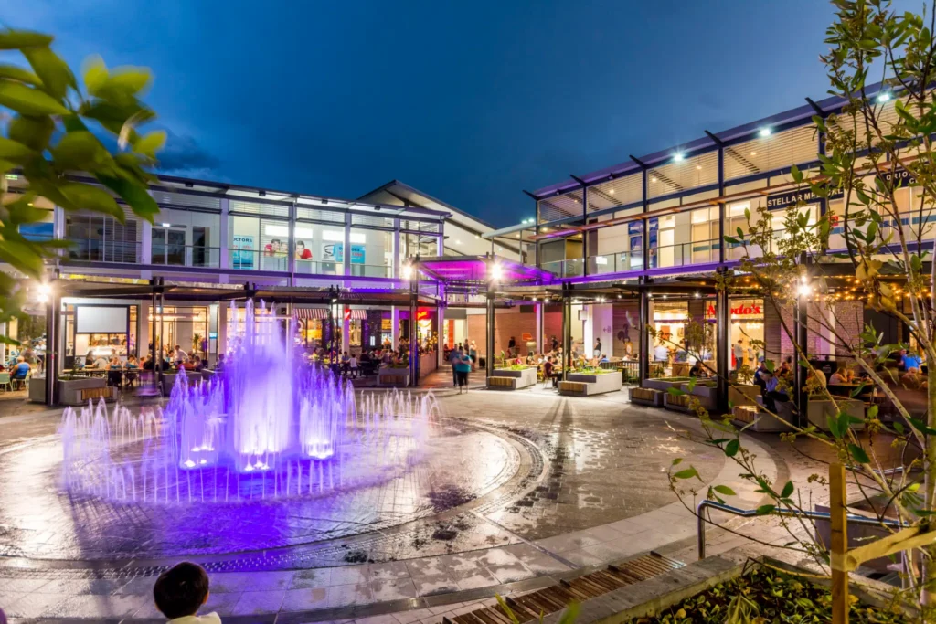 A vibrant outdoor shopping center at dusk with a glowing purple-lit fountain in the foreground and people dining at restaurants and cafes around the courtyard. The modern buildings are illuminated, creating a lively atmosphere.
