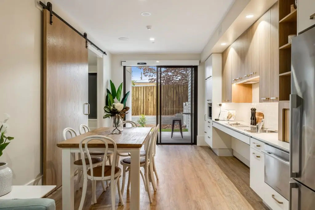 Modern kitchen and dining area with wood flooring, light wood cabinets, a wooden dining table with four chairs, sliding barn door, and glass doors leading to an outdoor patio with trees and a wooden fence.