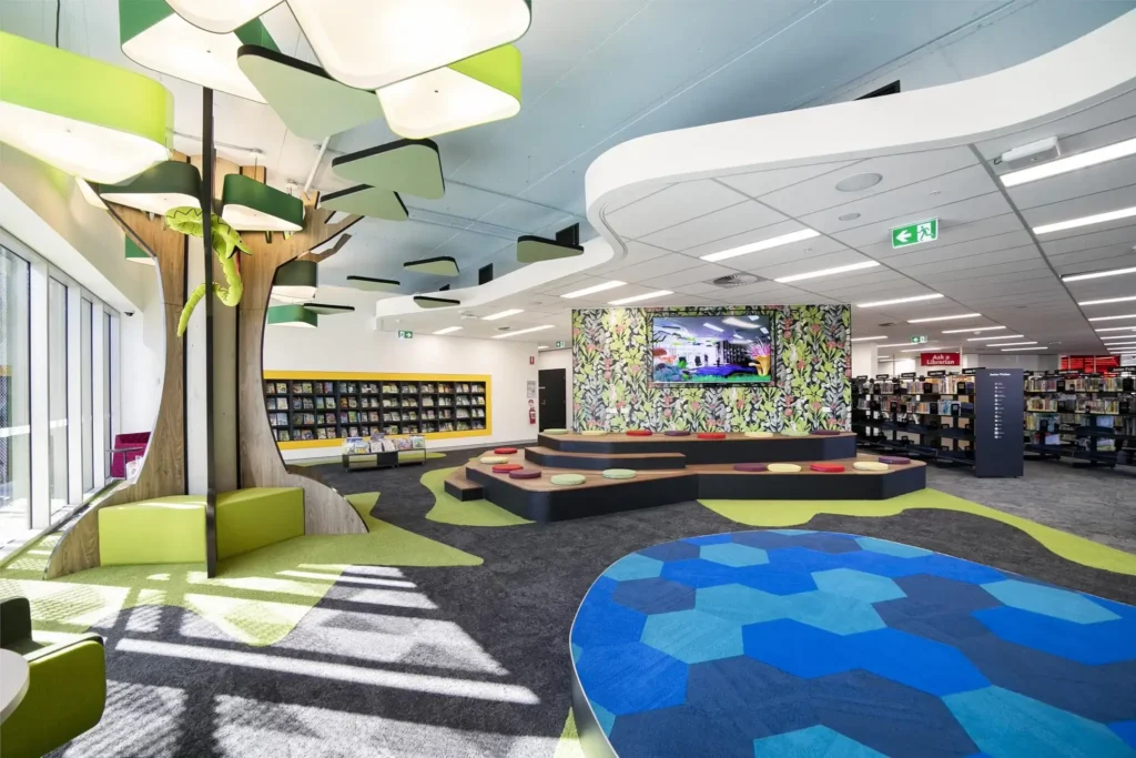 A modern library children’s area with colorful seating, a tree-shaped structure, bookshelves, a patterned wall with a large screen, and bright geometric carpet designs. Natural light fills the space.