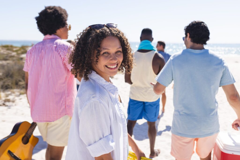 A woman smiling at the camera stands with friends on a sunny beach. The group is casually dressed, and one person holds a guitar. The ocean and blue sky are visible in the background.
