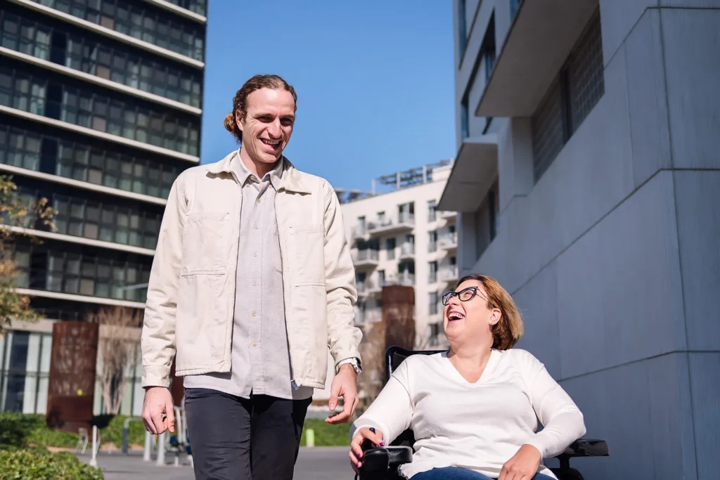 A man walks beside a woman in a wheelchair outdoors. Both are smiling and enjoying a sunny day, surrounded by modern buildings.
