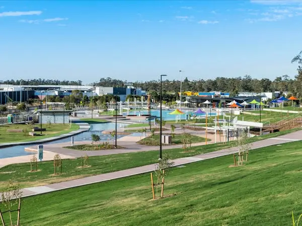 A modern park with green lawns, young trees, pathways, shaded seating areas, and buildings in the background under a blue sky with scattered clouds.