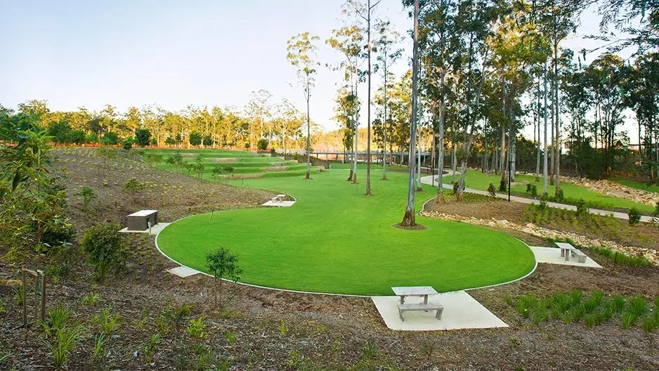 A landscaped park with green lawns, scattered trees, paved pathways, and several concrete picnic tables with benches. The area is surrounded by native plants and forested areas under a clear sky.