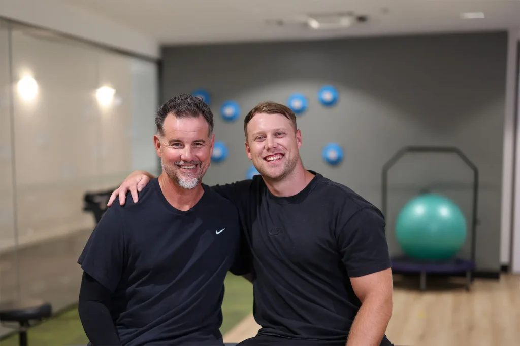 Two men in athletic wear sit closely together, smiling with arms around each other in a gym setting. Blue exercise balls and gym equipment are visible in the blurred background.