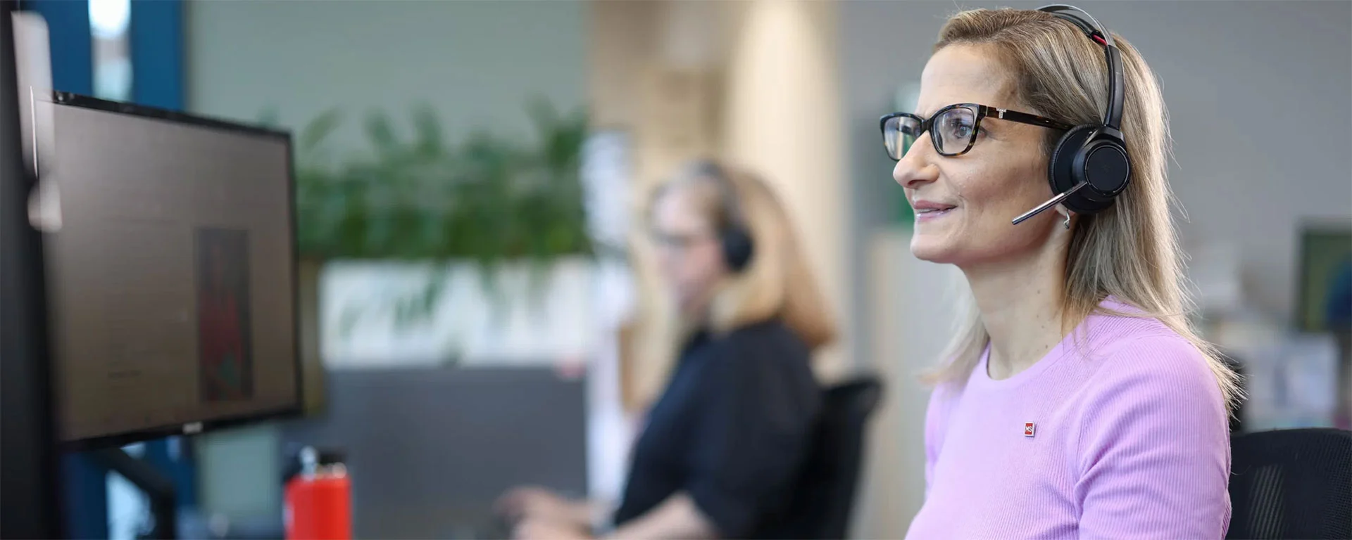 A woman wearing glasses and a headset smiles while working at a computer in an office. Another person in the background also works at a computer. The setting appears professional and modern.