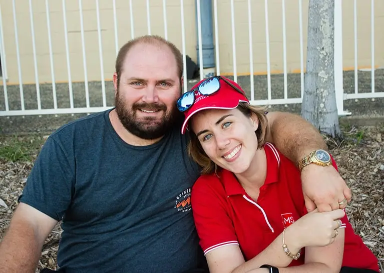 A man with a beard in a dark t-shirt sits next to a smiling woman in a red polo shirt and red cap. He has his arm around her shoulders, and they are sitting outdoors by a white metal fence.