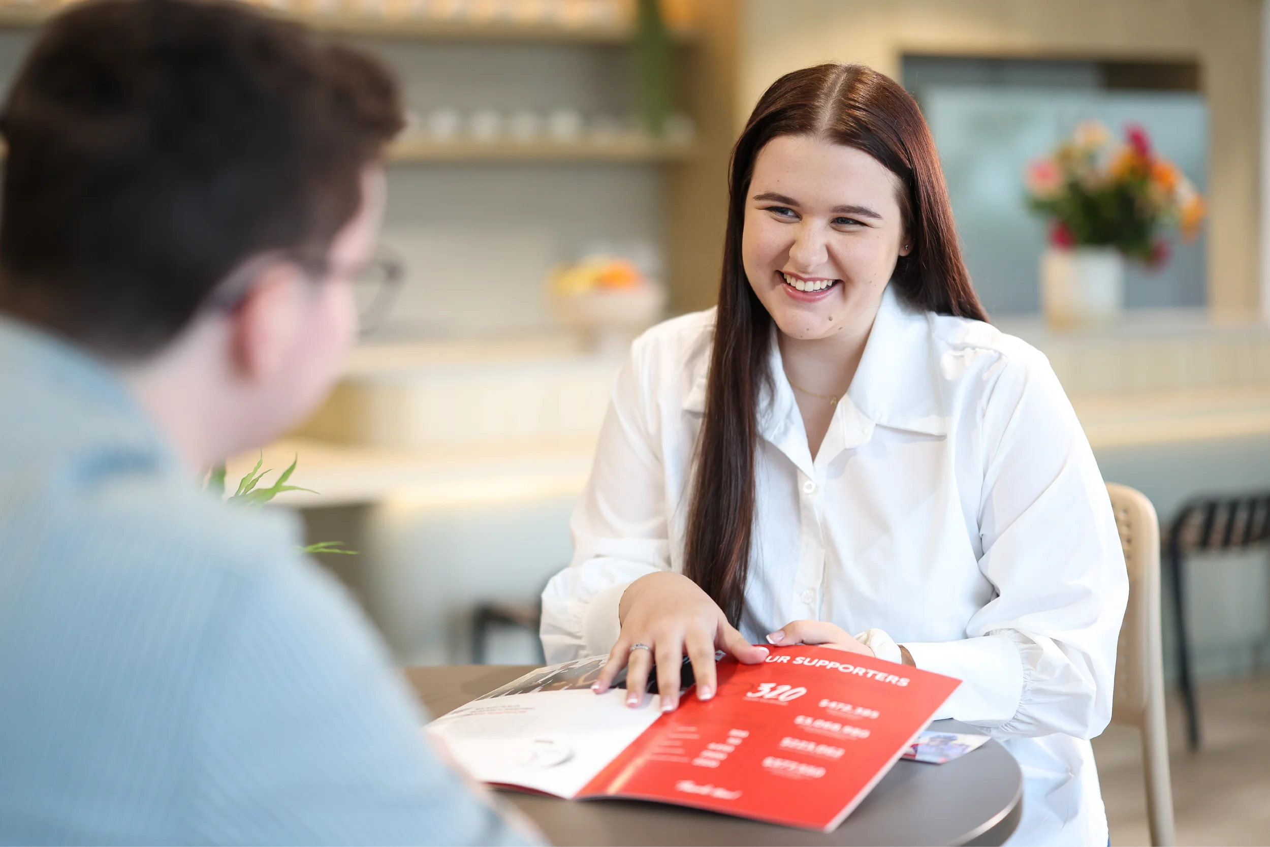 A woman with long brown hair, wearing a white shirt, smiles while talking to a man across a table. She is holding a red brochure. The background is a bright, modern indoor setting with shelves and flowers visible.