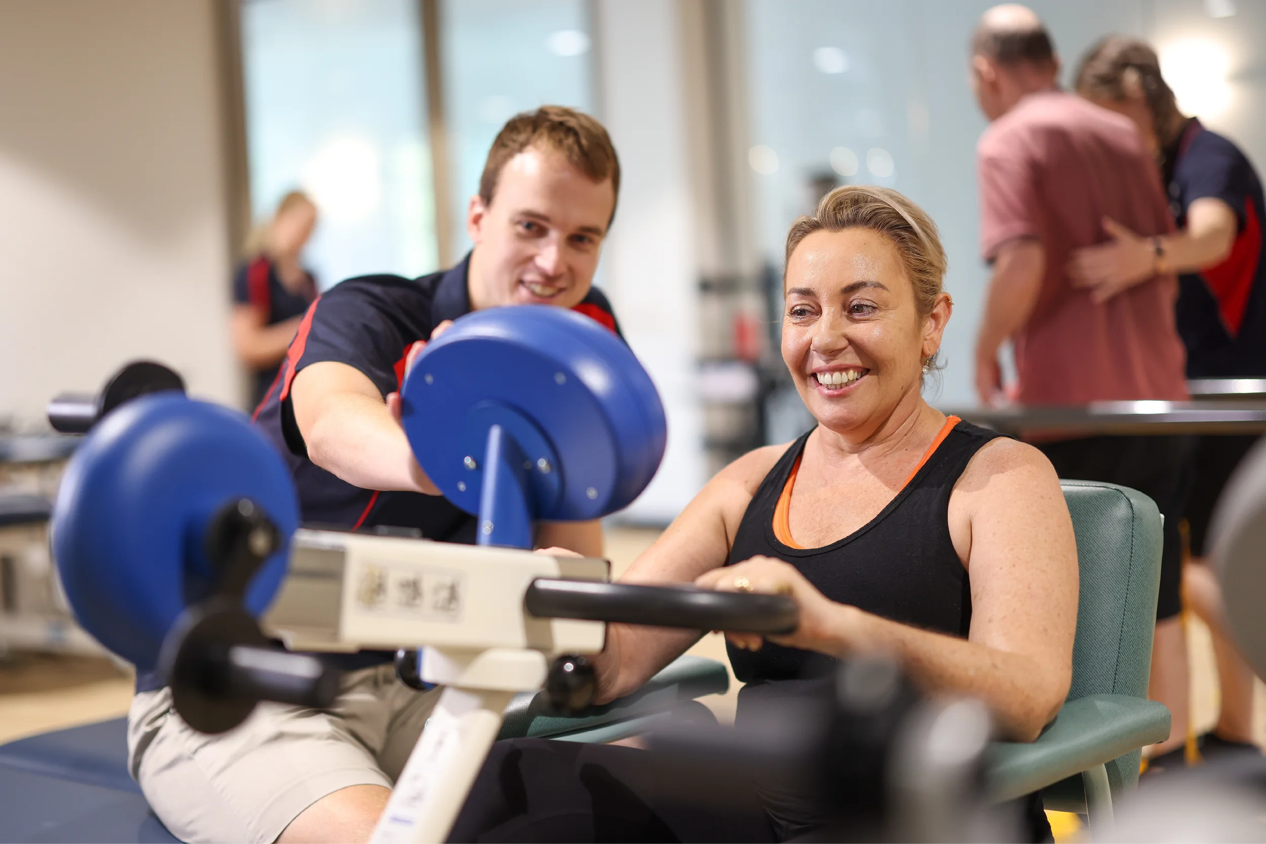 A smiling woman uses a seated exercise machine while a man beside her provides guidance. Both are in a bright gym or rehabilitation center with other people visible in the background.