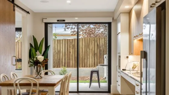 Modern kitchen and dining area with a wooden table and chairs, large glass sliding doors leading to a fenced backyard, and natural light illuminating the space. Plants and simple decor add a cozy touch.