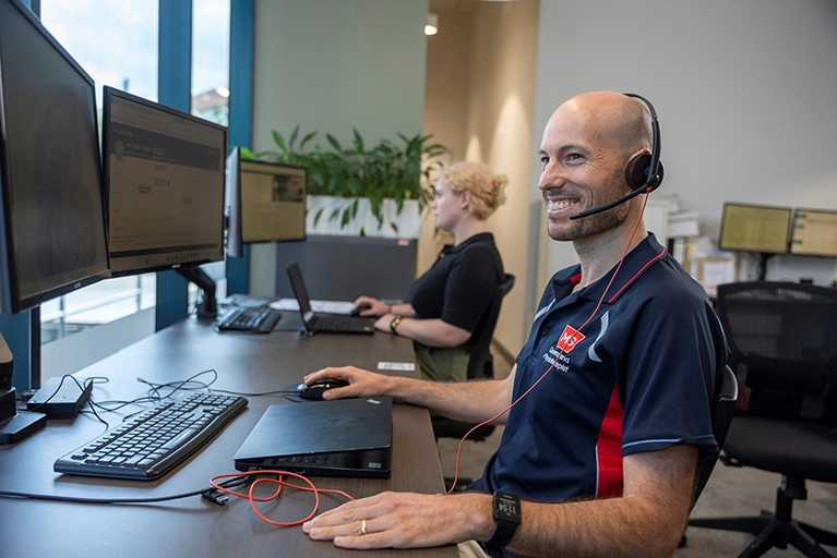 Two people work at desks with computers and headsets in a modern office. The man in the foreground smiles at his monitor, while a woman works in the background. Large windows and plants are visible. 