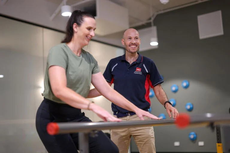 A woman practices walking at parallel bars in a rehab center while a smiling therapist in a navy blue polo shirt guides and encourages her.