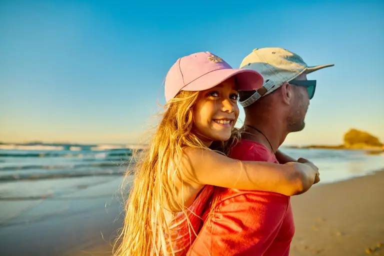 A smiling young girl wearing a pink cap gets a piggyback ride from a man in a light hat and sunglasses on a sandy beach at sunset, with waves and blue sky in the background.