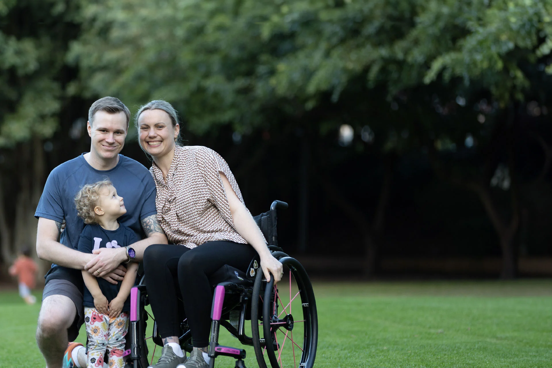 A smiling family sits together on grass in a park. An adult woman in a wheelchair is beside an adult man, and a young child stands in front, looking up at the adults. Trees fill the background.