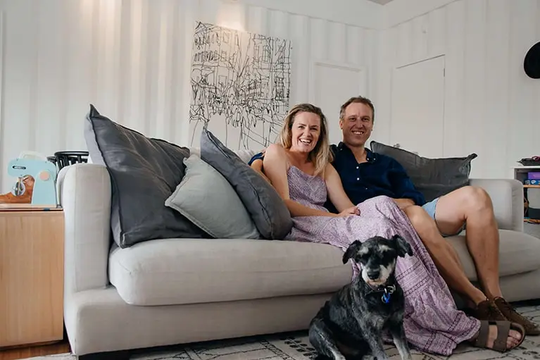 A smiling couple sits close together on a light-colored sofa in a cozy living room, with a small black and gray dog sitting on the floor in front of them. The room has neutral decor and wall art behind the couch.