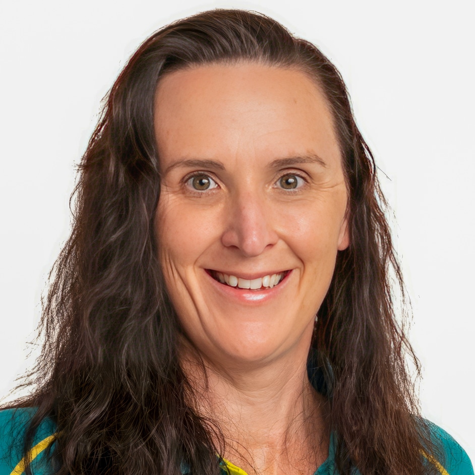 A woman with long wavy dark brown hair smiles at the camera. She is wearing a teal shirt with yellow accents and is posed against a plain white background.