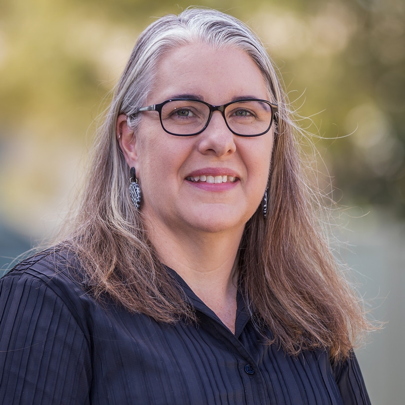 A woman with long, straight gray hair and glasses smiles outdoors. She is wearing a black blouse and earrings, with a blurred, sunlit background of greenery.