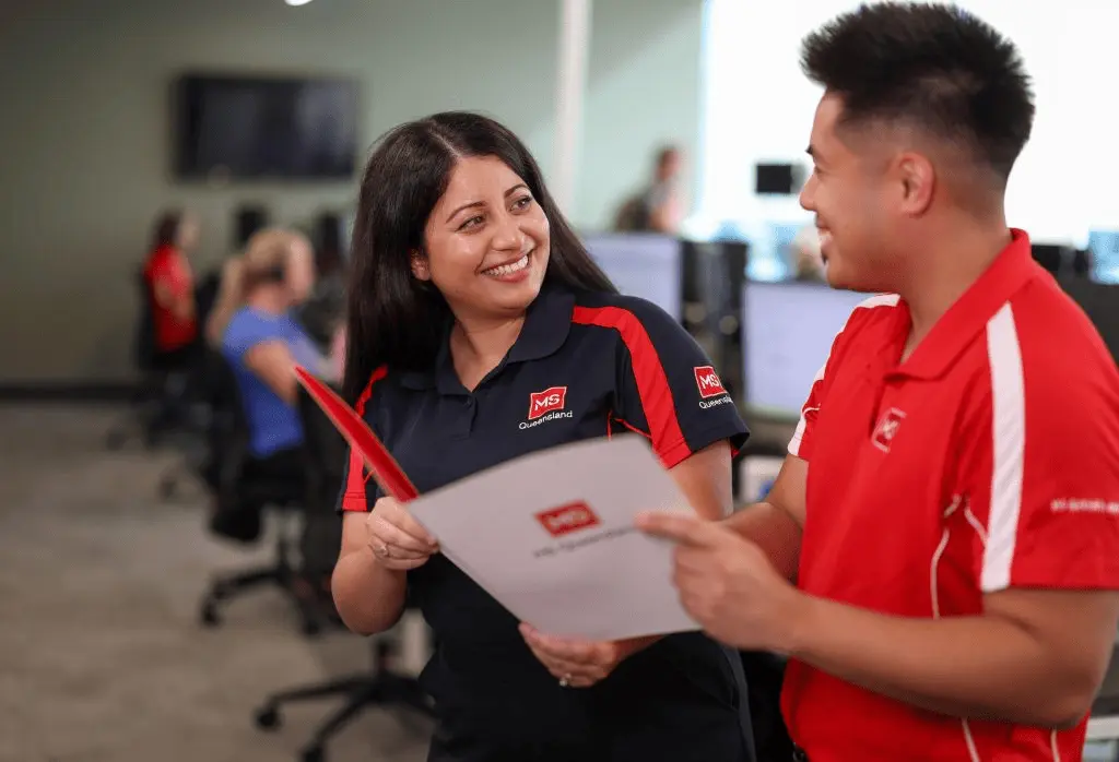 Two coworkers in red and navy MS Queensland uniforms smile and talk while holding documents in a modern office with other people working at computers in the background.