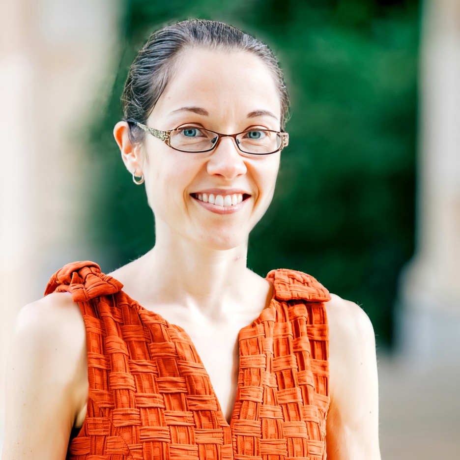 A woman with glasses and pulled-back hair smiles at the camera, wearing a textured orange sleeveless top. The background is blurred greenery and architecture.