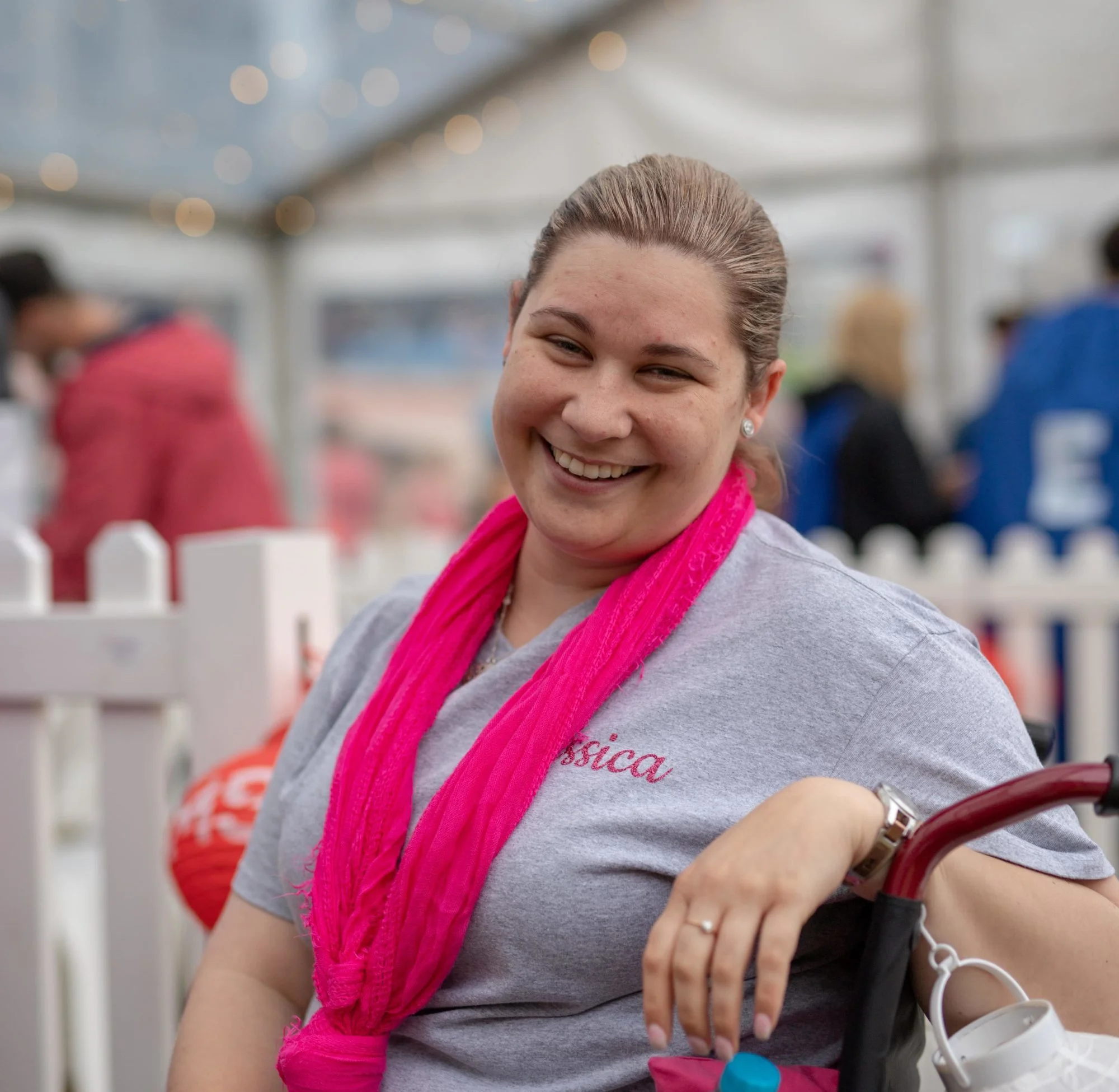 A woman with light brown hair tied back, wearing a gray shirt with "Jessica" on it and a bright pink scarf, smiles while sitting near a white picket fence at an outdoor event.