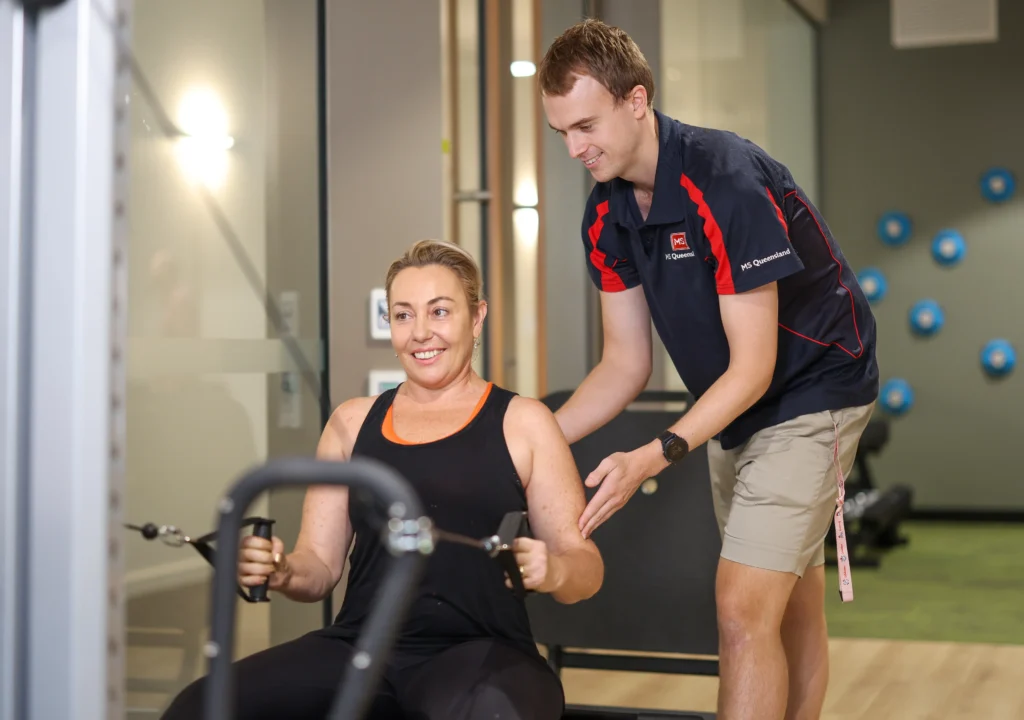A woman in workout clothes uses a rowing machine while a male trainer in a navy shirt and shorts stands beside her, offering guidance and encouragement in a gym setting.