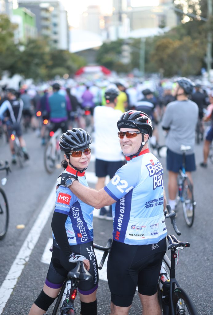 Two cyclists in matching blue jerseys stand together, smiling at the camera, surrounded by a large group of cyclists on a city street preparing for a cycling event. The city skyline is blurred in the background.